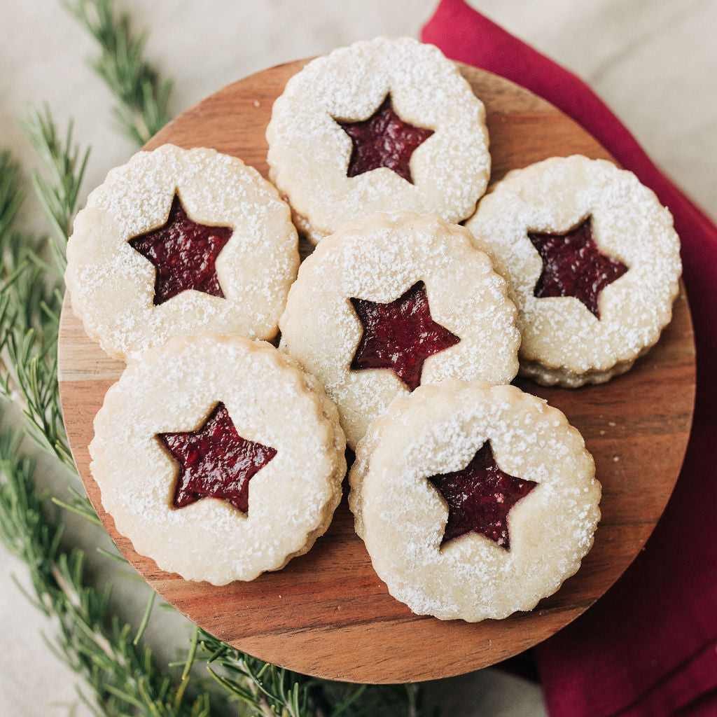 Decorative cookies with red star-shaped centers on a wooden board, surrounded by greenery and a red cloth.